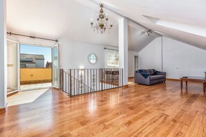 A spacious loft with a wooden floor, a grey sofa, a chandelier, and a view of a building outside.