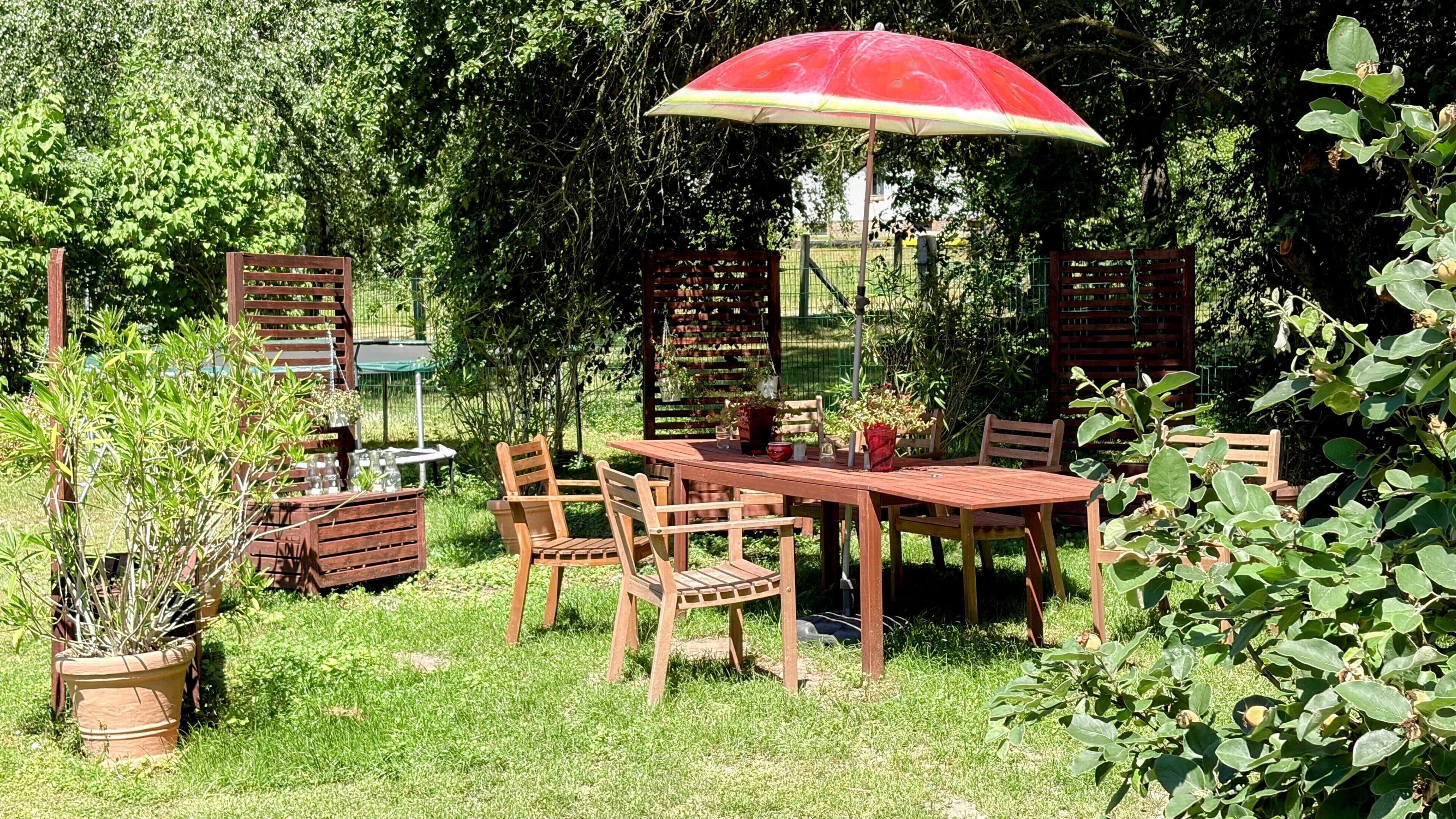 An outdoor patio with a wooden table and chairs, surrounded by greenery and a red umbrella.