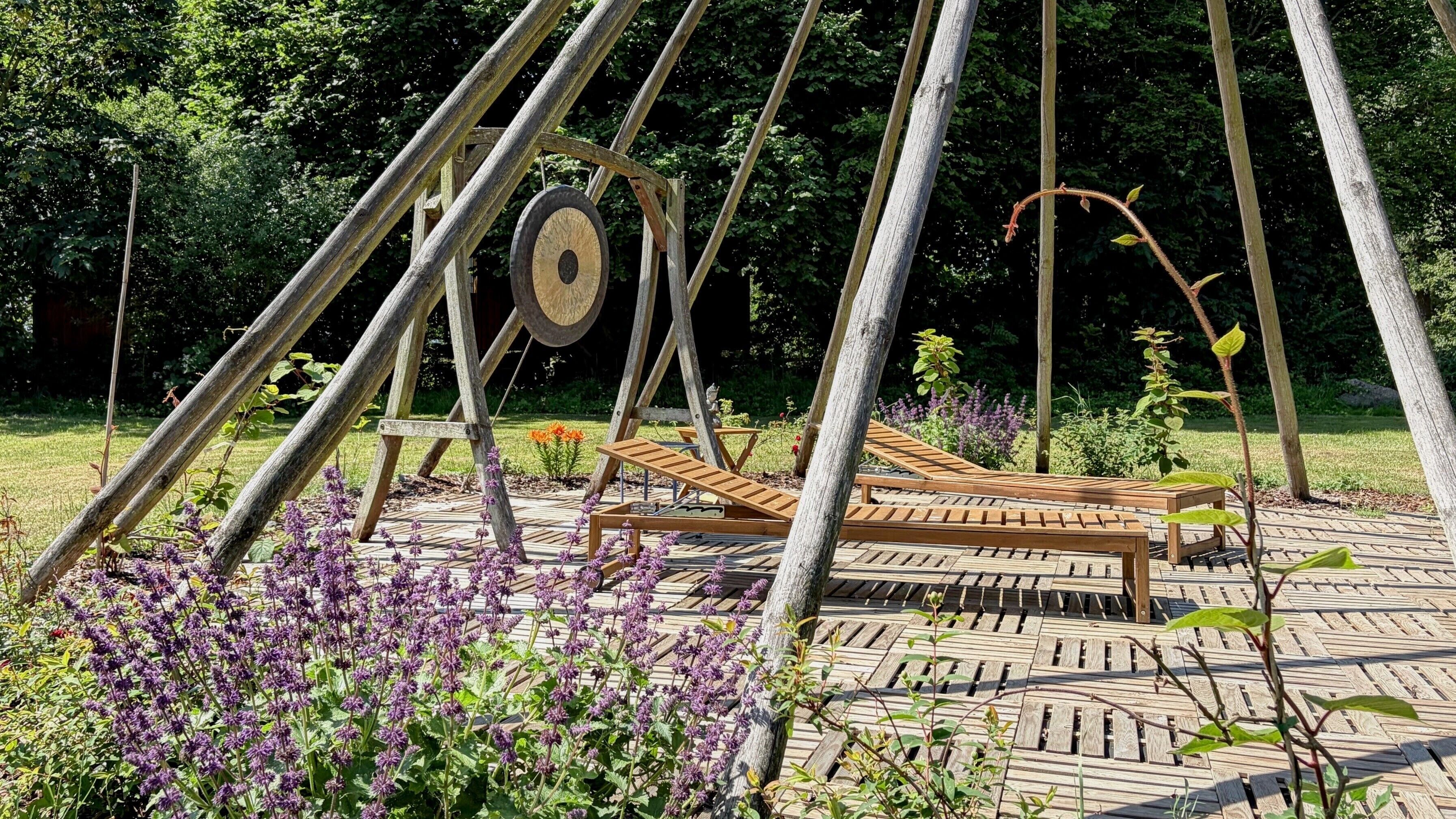 A wooden swing set with a bench, surrounded by a garden with purple flowers and a paved area.