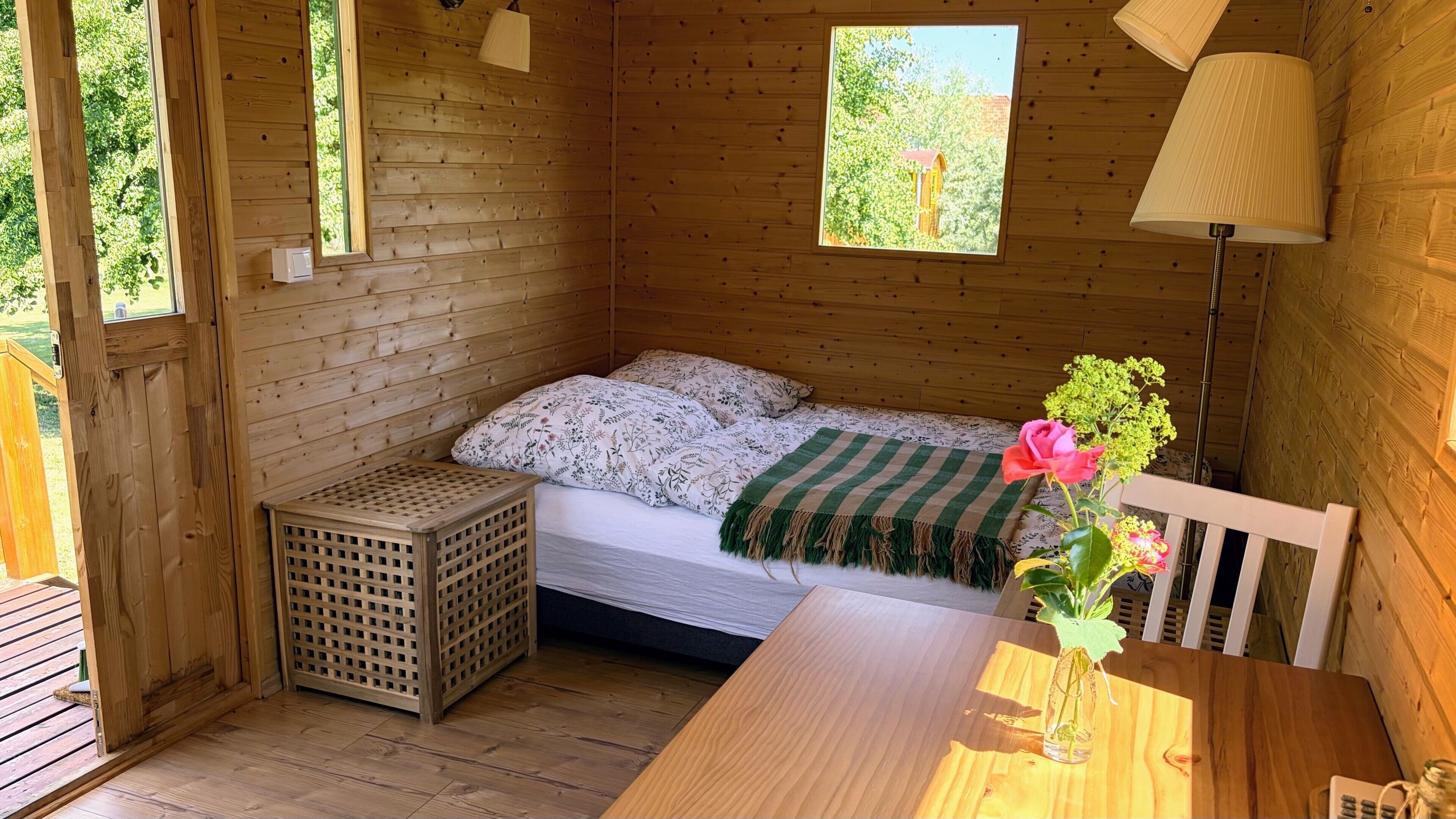 Ein Zimmer in einer Holzhütte mit einem Bett, einem Tisch, einer Lampe und einem Fenster.