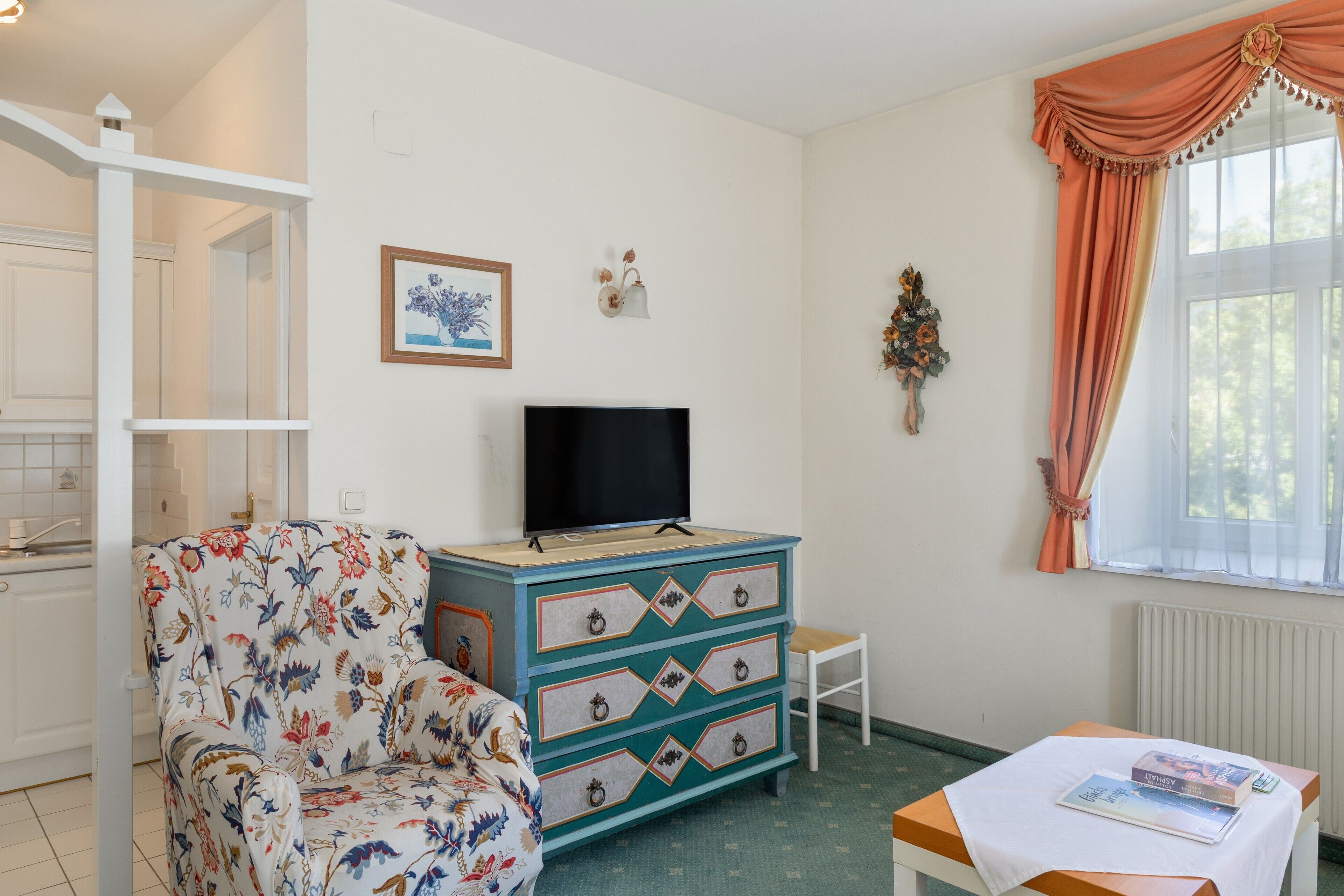 Une pièce avec une télévision posée sur une commode, une chaise à motifs floraux, une petite table avec des livres et une fenêtre avec des rideaux.