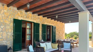 A stone exterior wall with green shutters, a round stone table with a glass top, and a wooden bench with cushions.