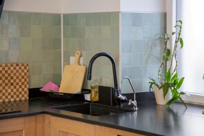 Une cuisine avec un évier noir, une planche à découper en bois, une bouteille de liquide vaisselle et une plante en pot.