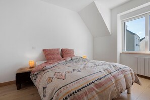 A bedroom with a sloped ceiling, a large window, a bed with patterned bedding, and two bedside lamps.