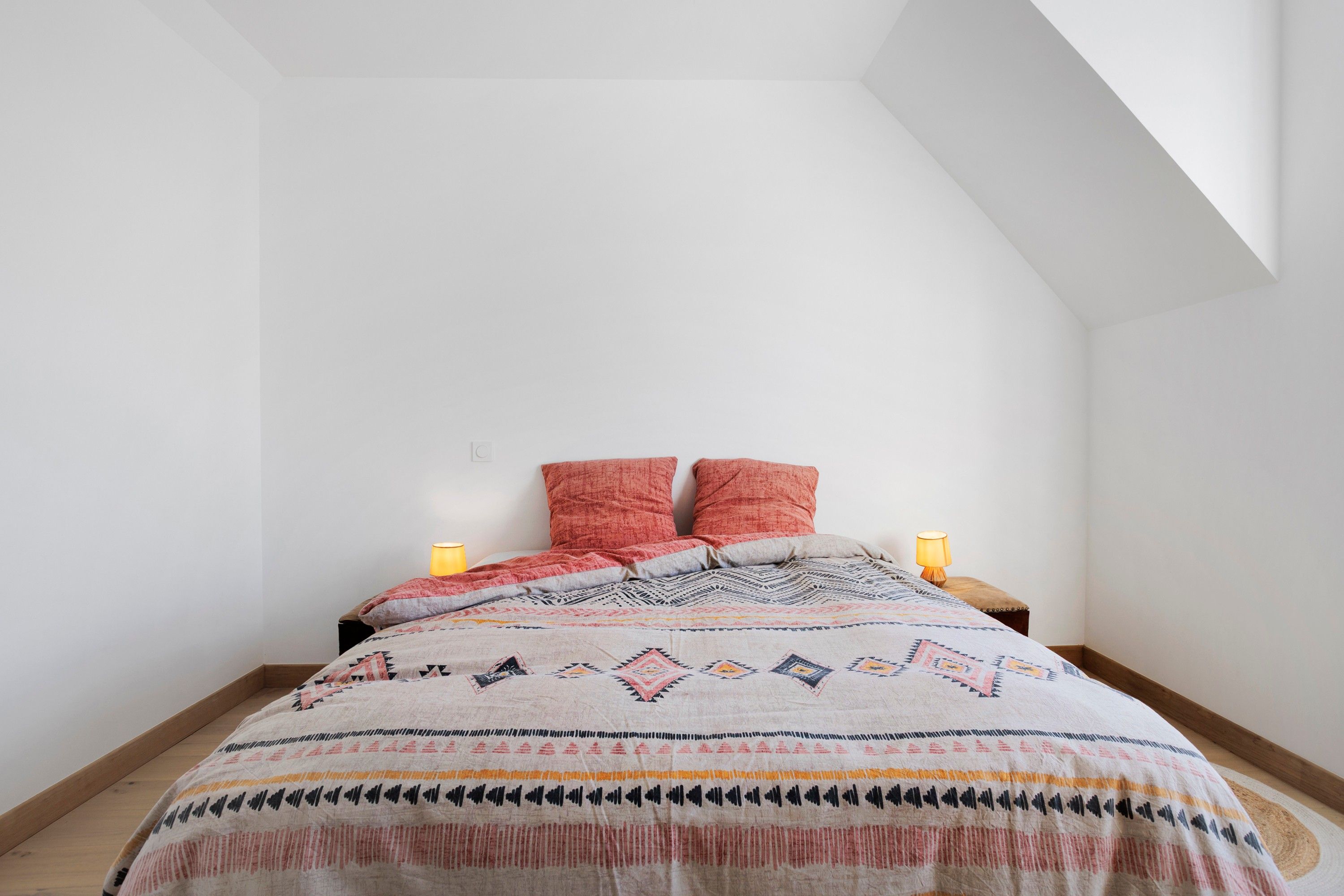 A bedroom with a sloped ceiling, a bed with a patterned bedspread, two pillows, two bedside lamps, and a wooden floor.