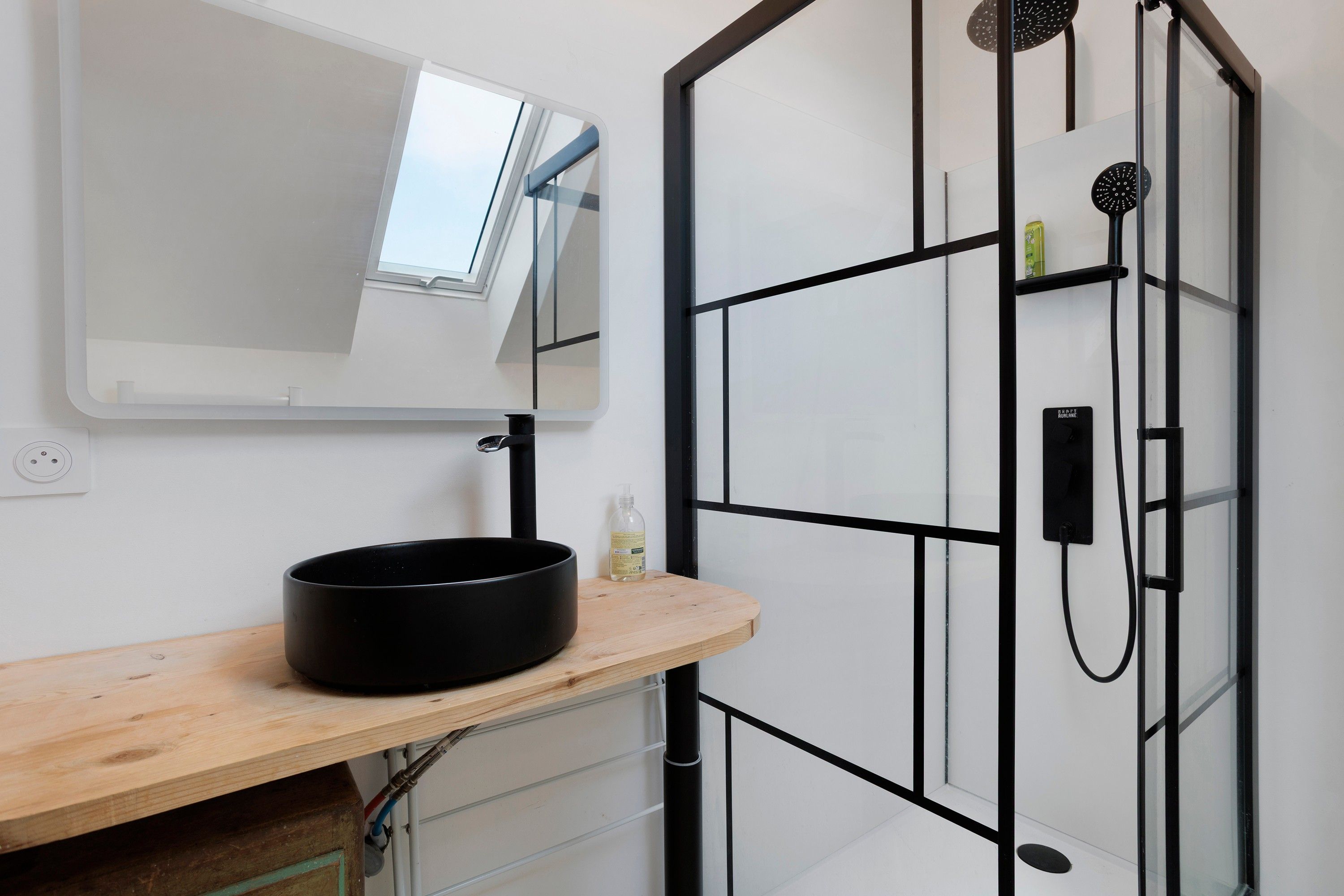 Un baño con cabina de ducha negra, un espejo, un lavabo con recipiente negro y un estante de madera.