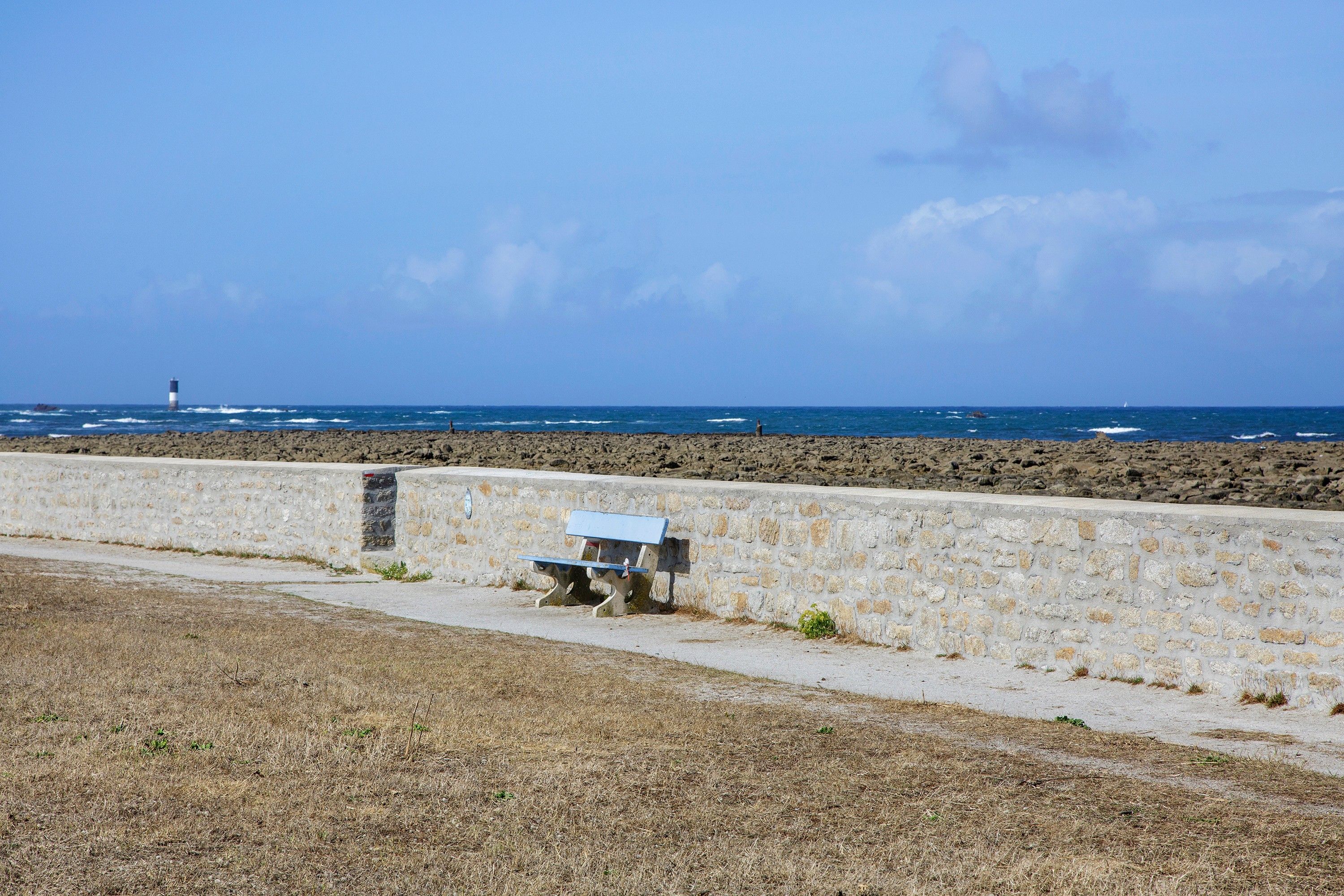 A coastal view with a stone wall, a bench, and a lighthouse in the distance.