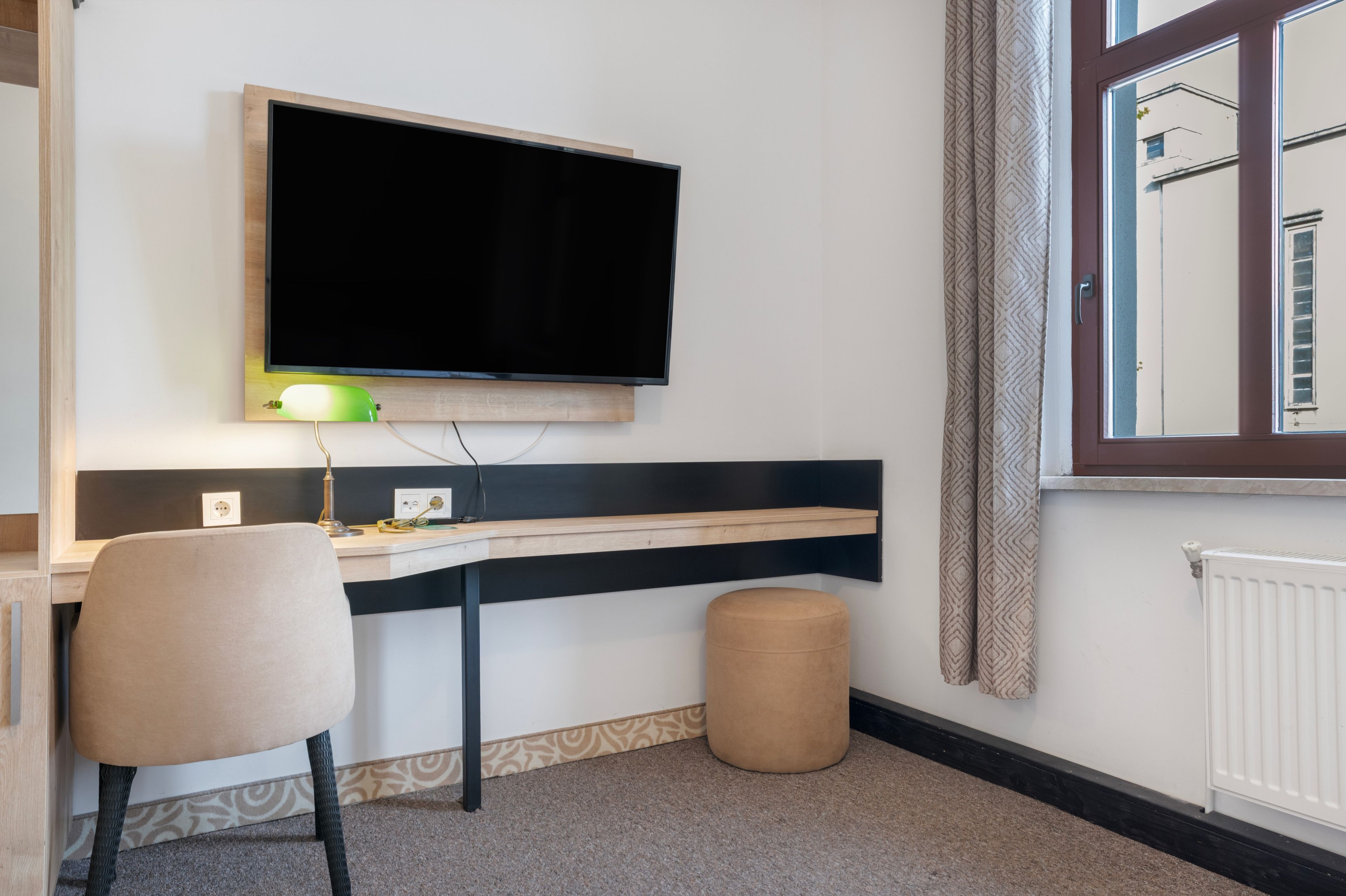 A room with a mounted flat-screen TV, a desk with a lamp, a chair, a small ottoman, and a window with curtains.
