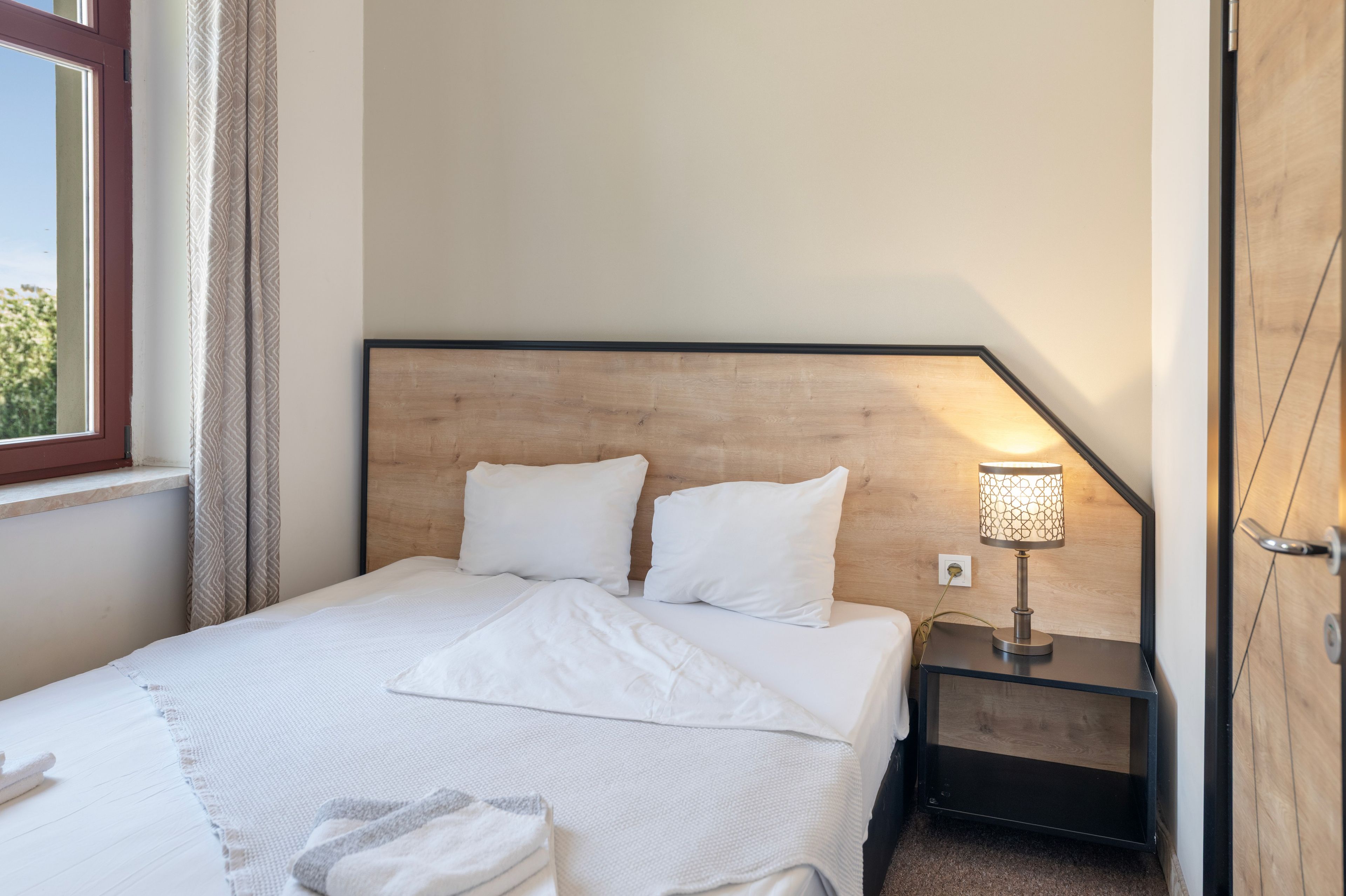 A neatly made bed with white linens, a bedside lamp, and a window with curtains.