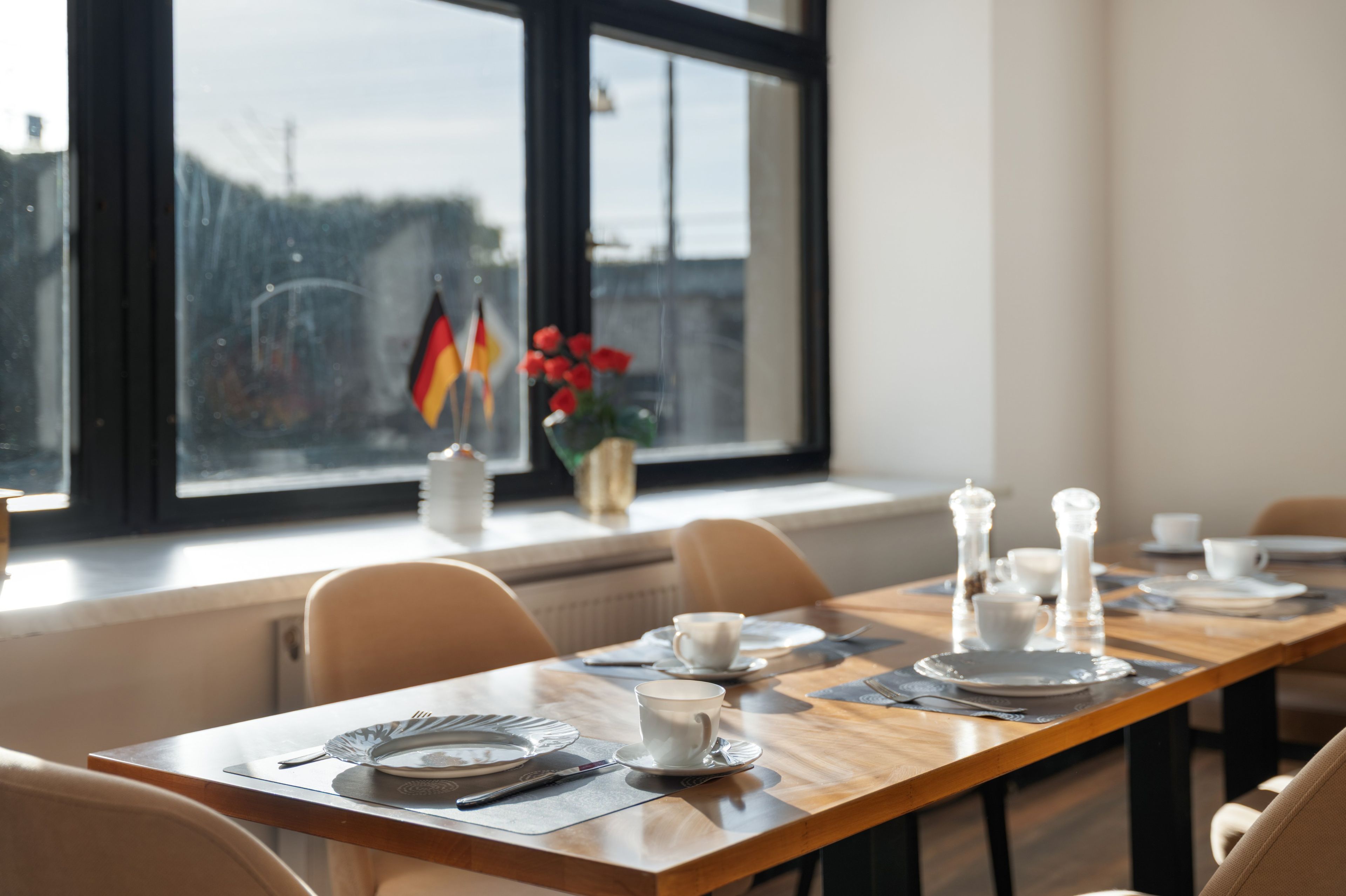 Comedor con una mesa dispuesta para dos, un florero con flores rojas y una ventana con banderas.