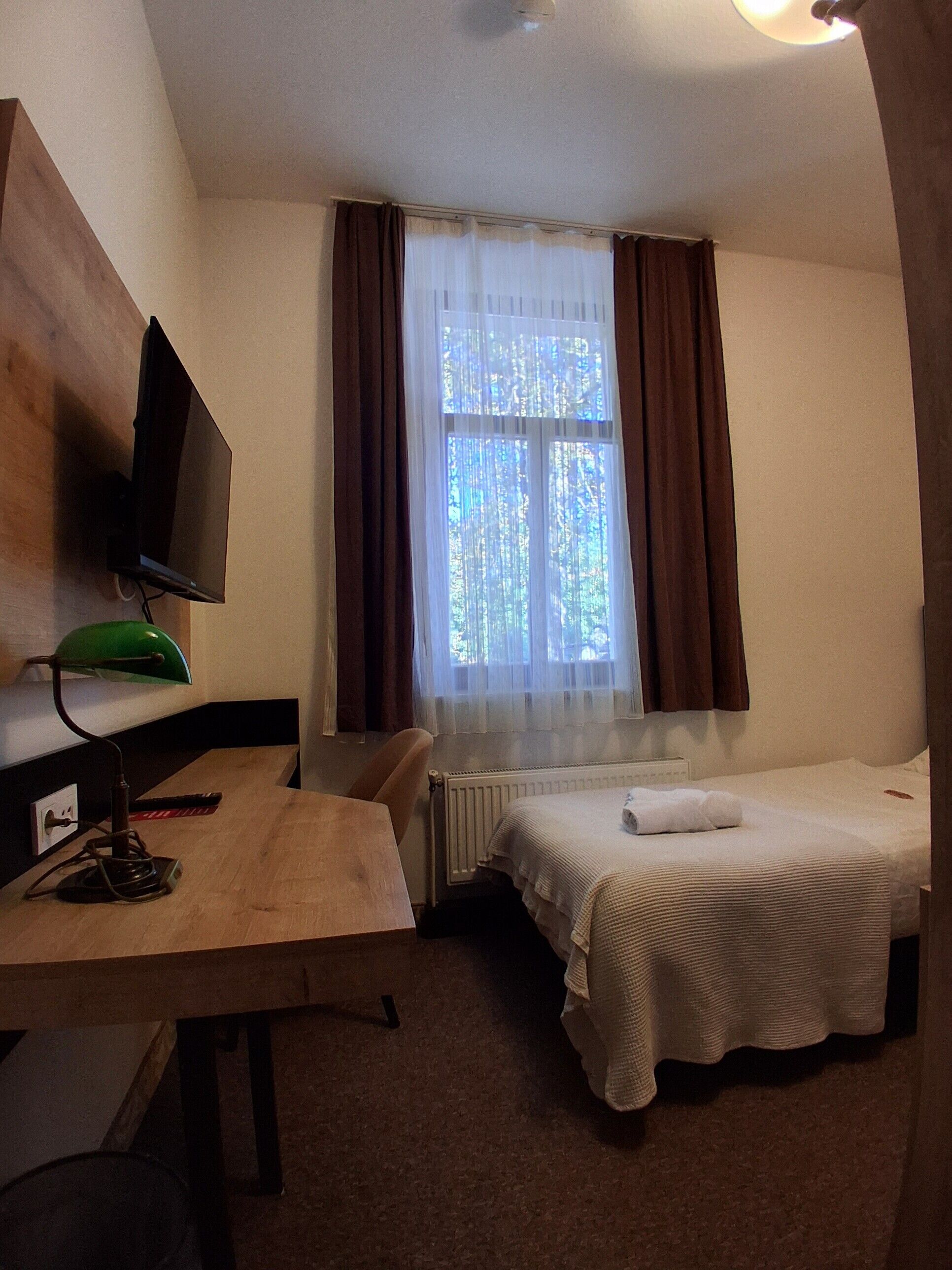 A hotel room with a single bed, a desk with a computer, a television, and a window with curtains.