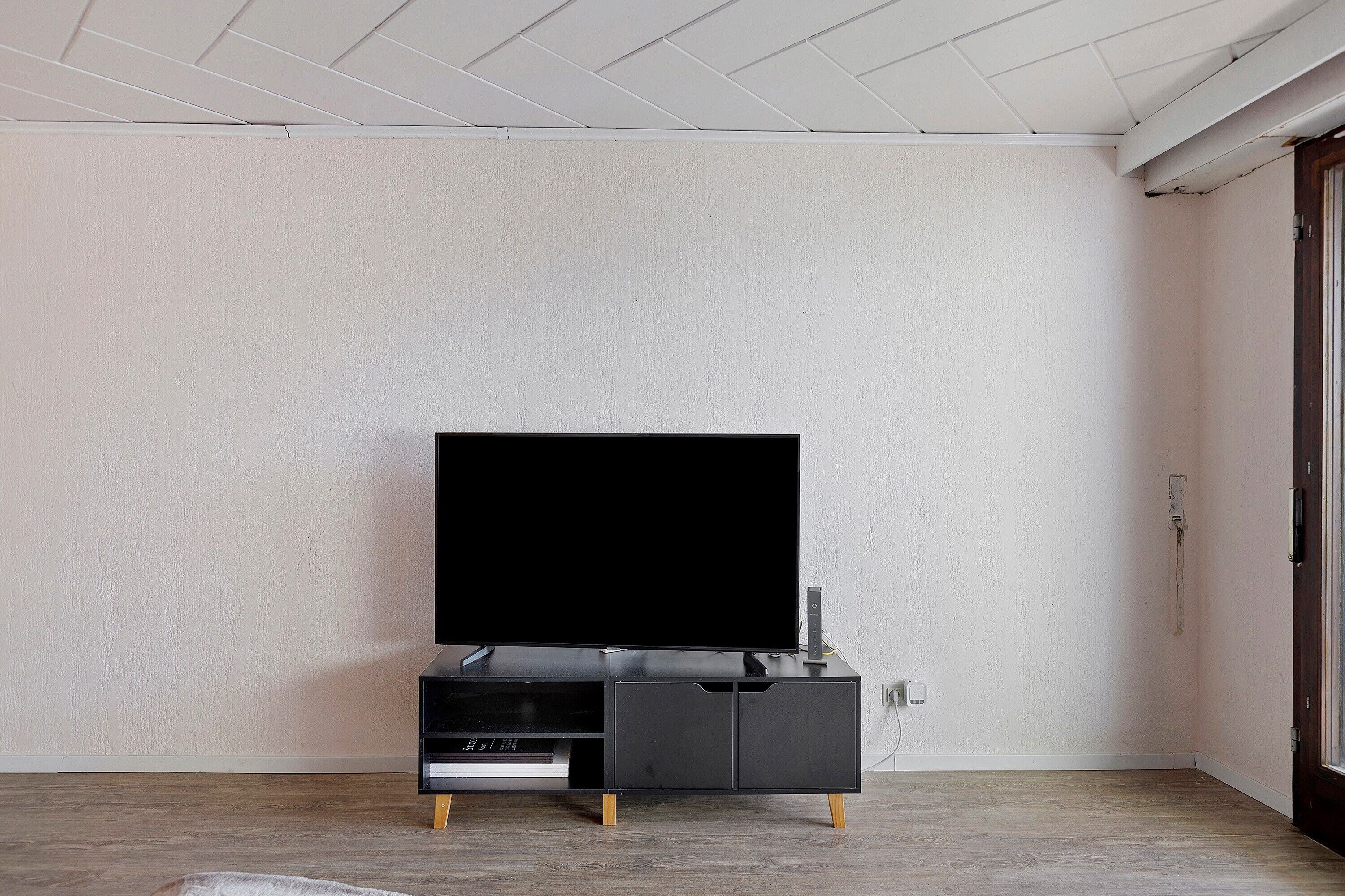 A minimalist room with a flat-screen TV on a black stand, a wooden floor, and a plain white wall.