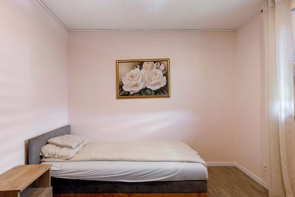 A bedroom with a single bed, a nightstand, a painting of roses, and a wooden bench.