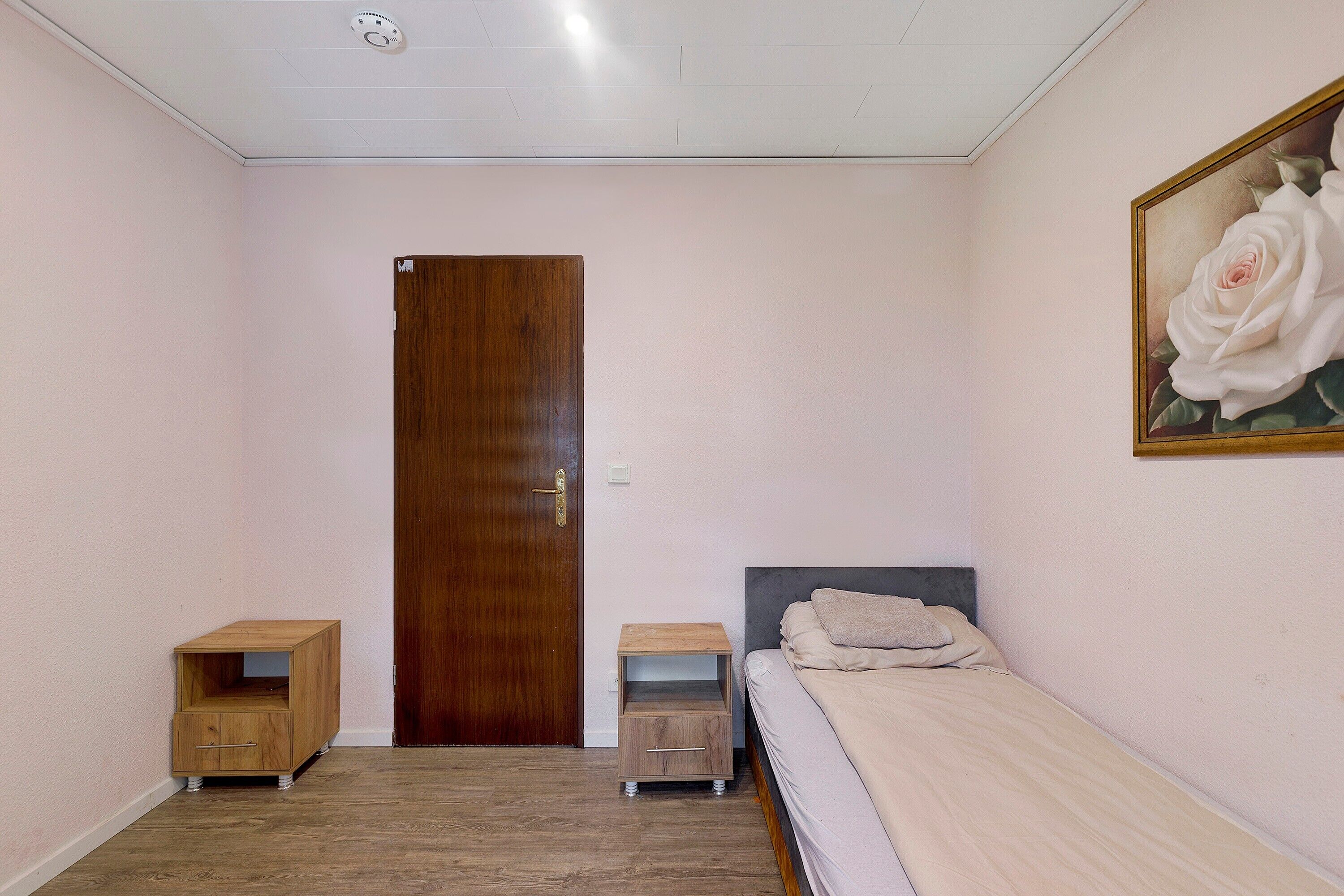 A bedroom with a wooden door, a bed with a grey headboard, a nightstand, a small wooden cabinet, and a painting of a white rose on the wall.