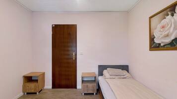 A bedroom with a wooden door, a bed with a grey headboard, a nightstand, a small wooden cabinet, and a painting of a white rose on the wall.