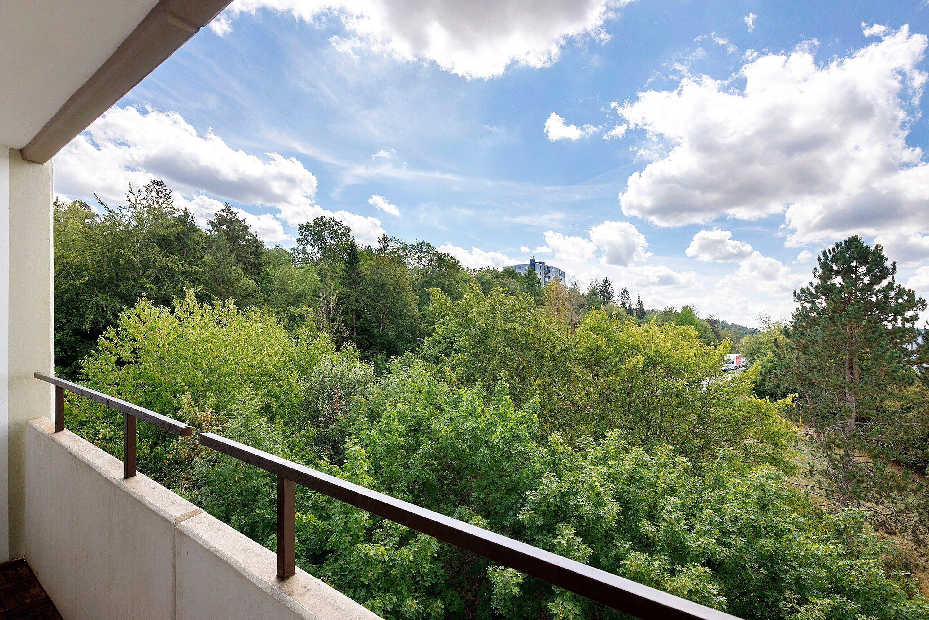 A balcony with a metal railing overlooking a dense forest and a building in the distance.