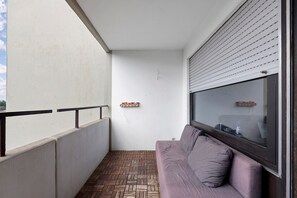 Un balcón con suelo de madera, pared blanca, ventana con persianas y sofá púrpura con cojines.
