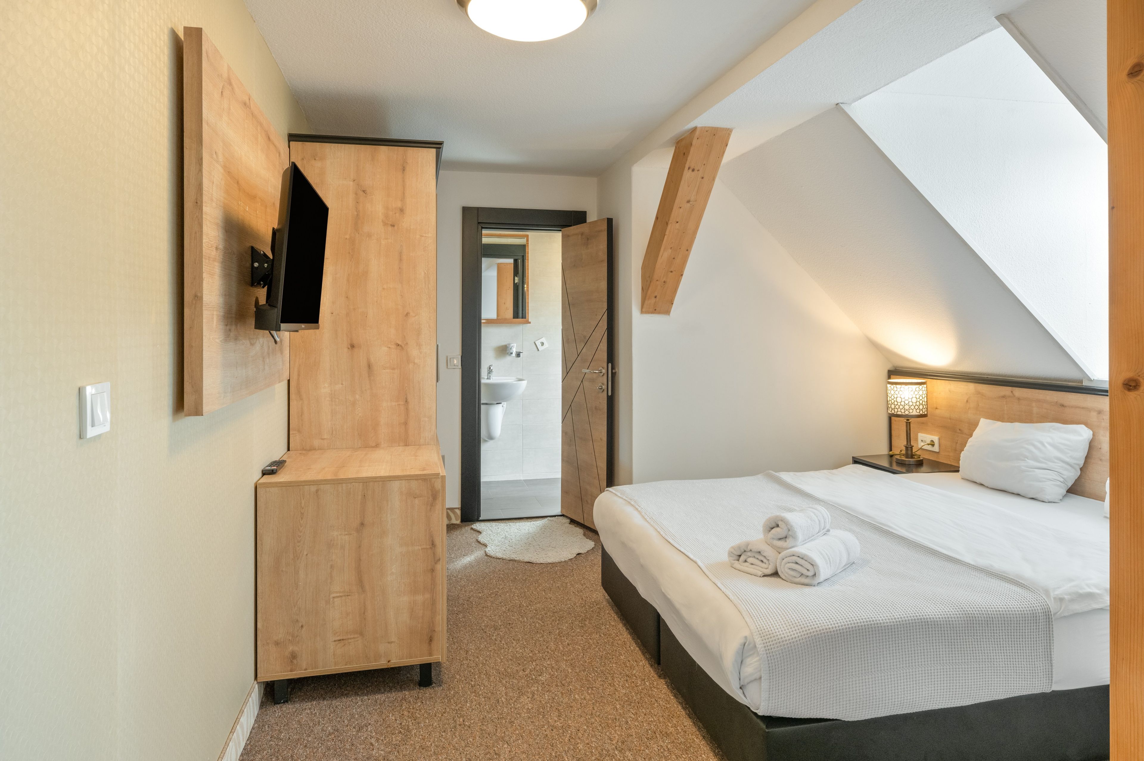 Ein modernes Hotelzimmer mit einem Bett, einem Holzschrank, einem an der Wand befestigten Fernseher und einem durch eine geöffnete Tür einsehbarer Badezimmer.