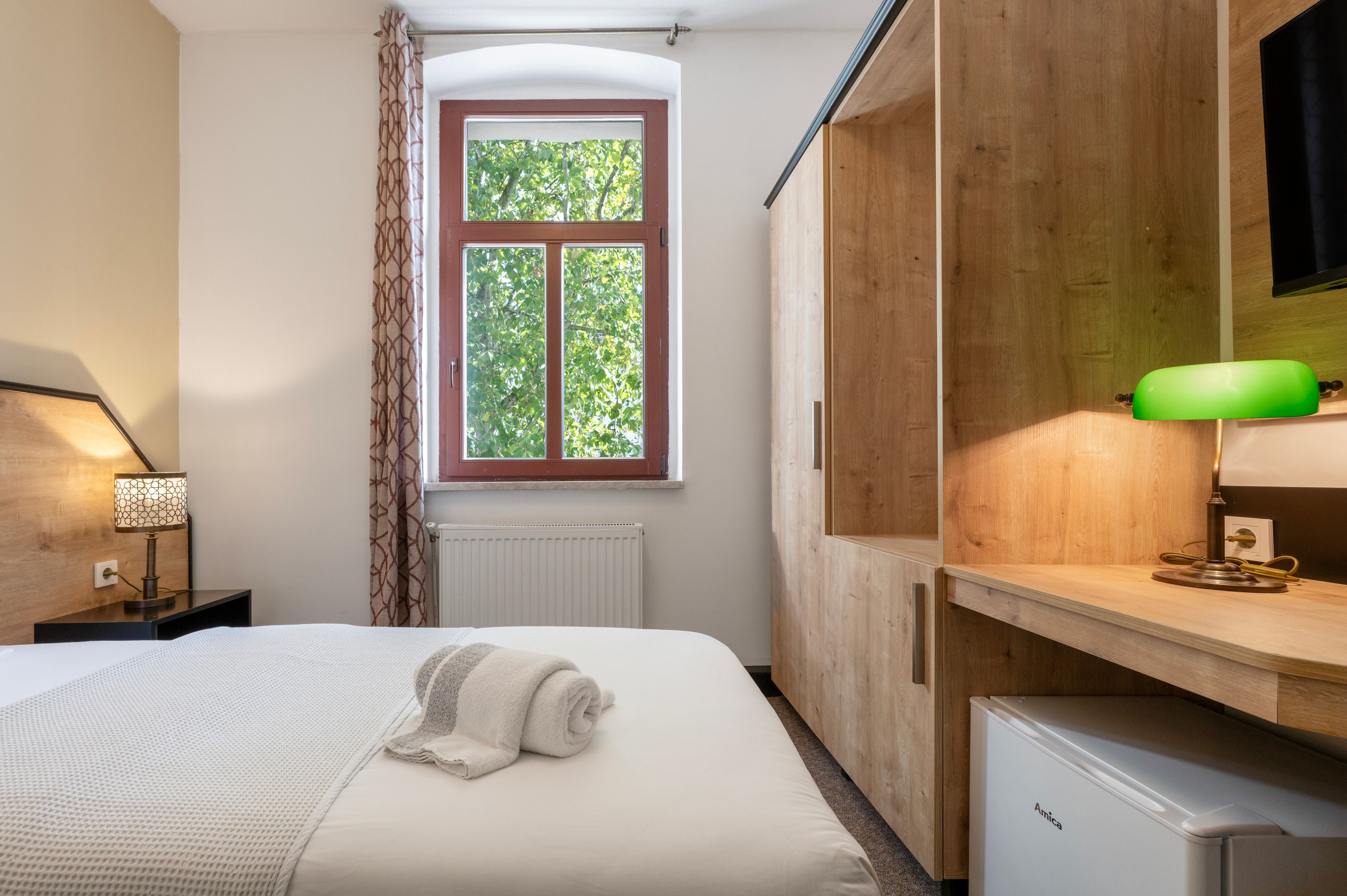 Ein Hotelzimmer mit Bett, Fenster mit Blick auf Bäume, Heizkörper, Holzschrank, Schreibtisch mit Lampe und einem kleinen Kühlschrank.