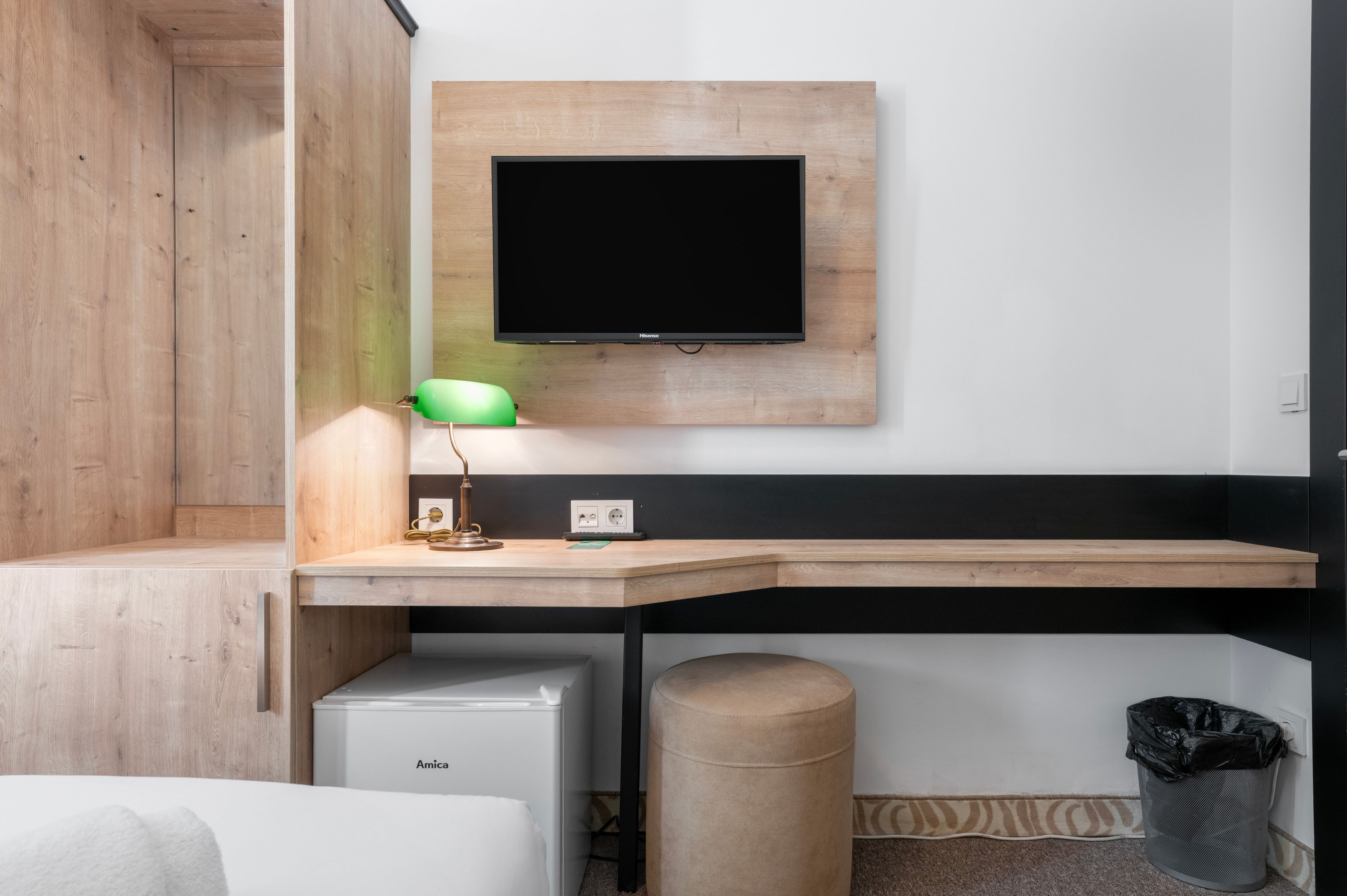 Ein modernes Hotelzimmer mit einem Holztisch, einem fest installierten Flachbildfernseher, einer grünen Schreibtischlampe, einem kleinen weißen Gerät und einem Mülleimer.