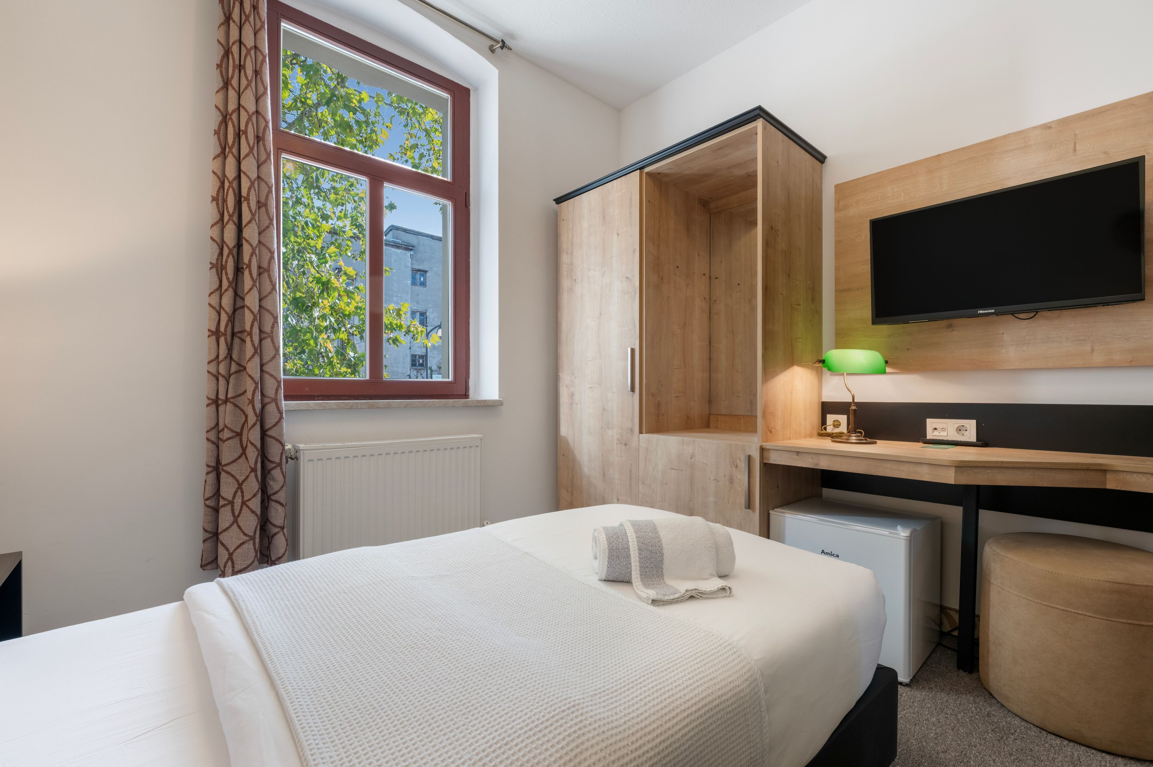 A hotel room with a bed, a desk with a television, a wardrobe, and a radiator.
