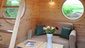 A cozy cabin interior with a loft bed, a sofa, a small table with a vase of flowers, and two windows offering views of greenery.