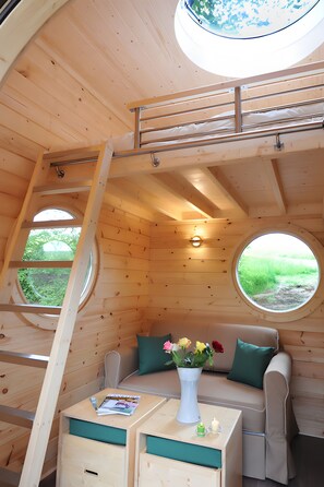 A cozy cabin interior with a loft bed, a sofa, a small table with a vase of flowers, and two windows offering views of greenery.