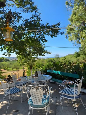 An outdoor patio with a round table and white metal chairs, surrounded by greenery and a clear view of the countryside. - Louisa's Cottage – Hilltop Retreat in Bochali (Zakynthos)