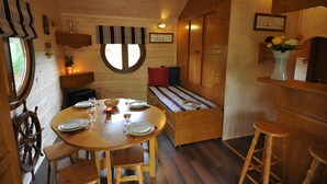 A cozy cabin interior with a wooden dining table set for four, a bench with pillows, and a small kitchen area with a sink and a vase of flowers.