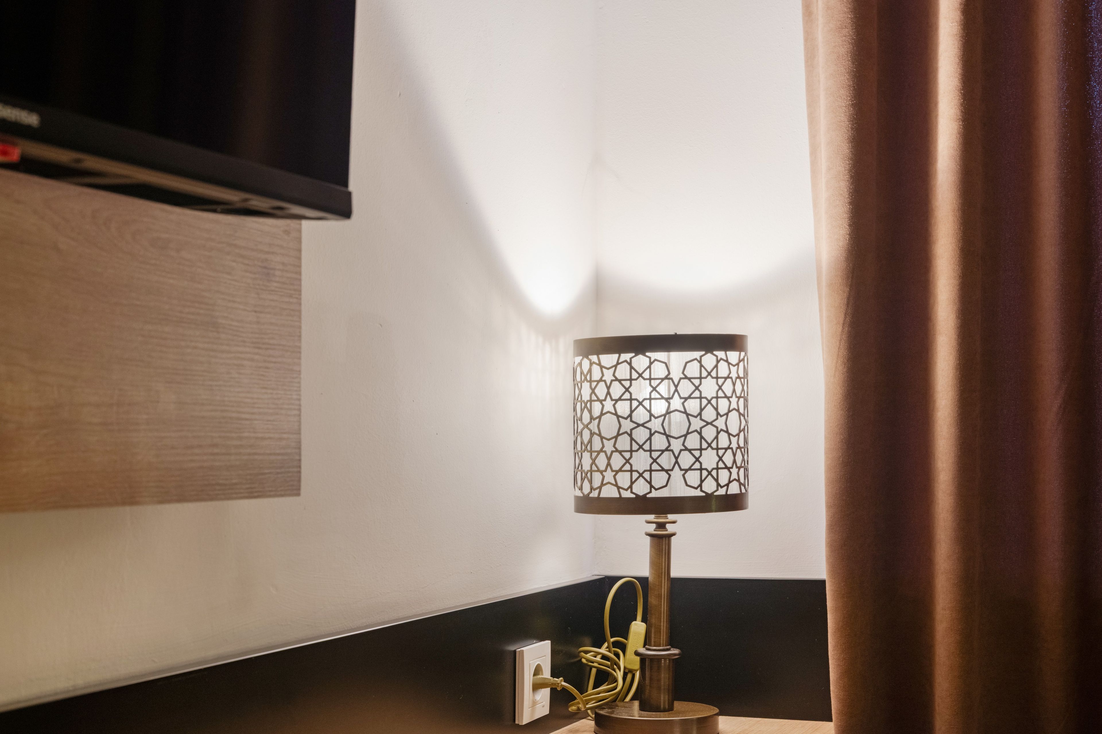 A corner of a room with a lamp on a wooden surface, a power outlet, and a wall-mounted television.