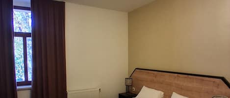 Habitación de hotel con cama, una silla, mesita de noche con lámpara, ventana con cortinas e iluminación en el techo.