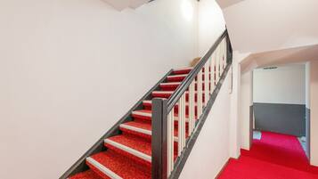 A staircase with red carpeted steps and a black handrail, leading to a room with a white wall and a red carpeted floor.