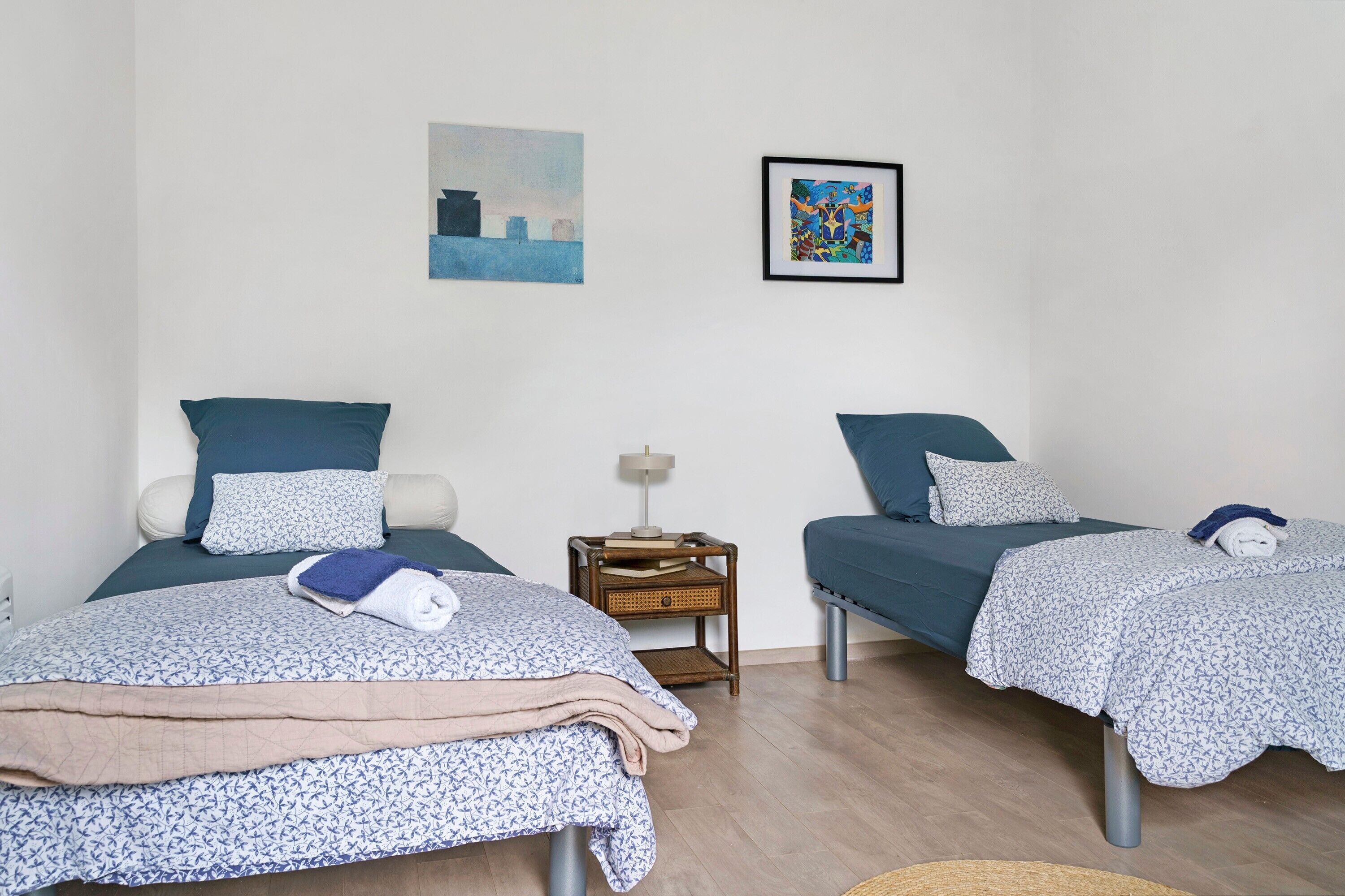 Een kamer met twee bedden, elk voorzien van een blauwe spreidek en kussens, een klein nachtkastje met een lamp, en twee kunstwerken in lijsten aan de muur.