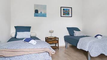 Een kamer met twee bedden, elk voorzien van een blauwe spreidek en kussens, een klein nachtkastje met een lamp, en twee kunstwerken in lijsten aan de muur.