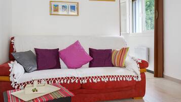 A red sofa with white and purple cushions, a small table with a placemat, and a window with a view of greenery.