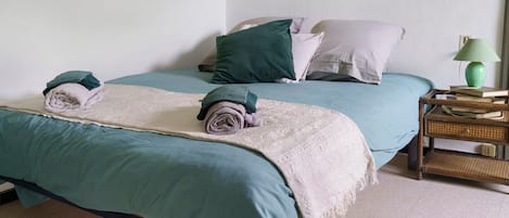 Un lit recouvert d’une couvre-lit turquoise, avec des serviettes enroulées et une couverture blanche. On y trouve une table de chevet avec une lampe et des livres.