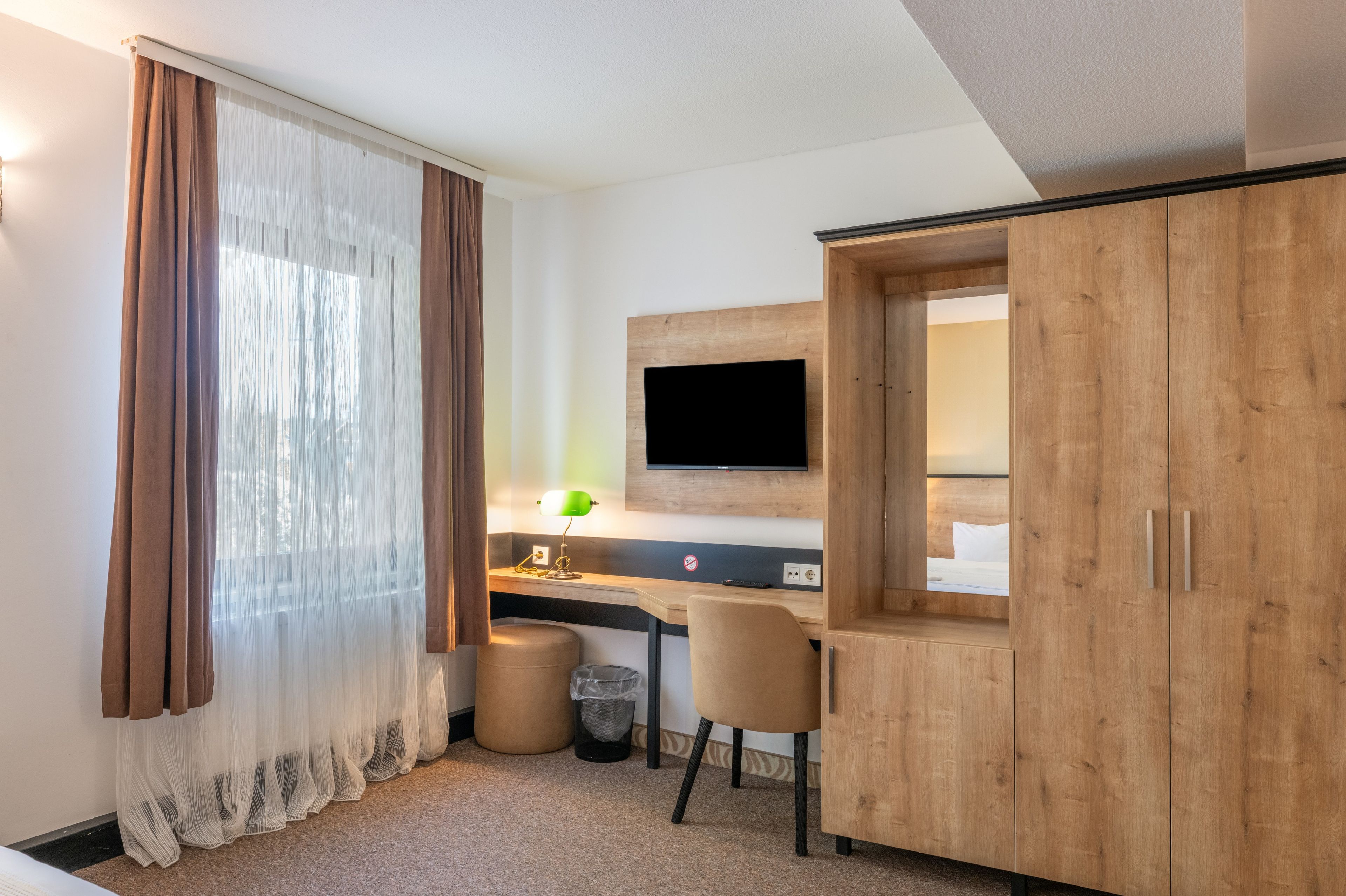 A hotel room with a wooden desk, a television mounted on the wall, a chair, a trash bin, and a window with sheer curtains.