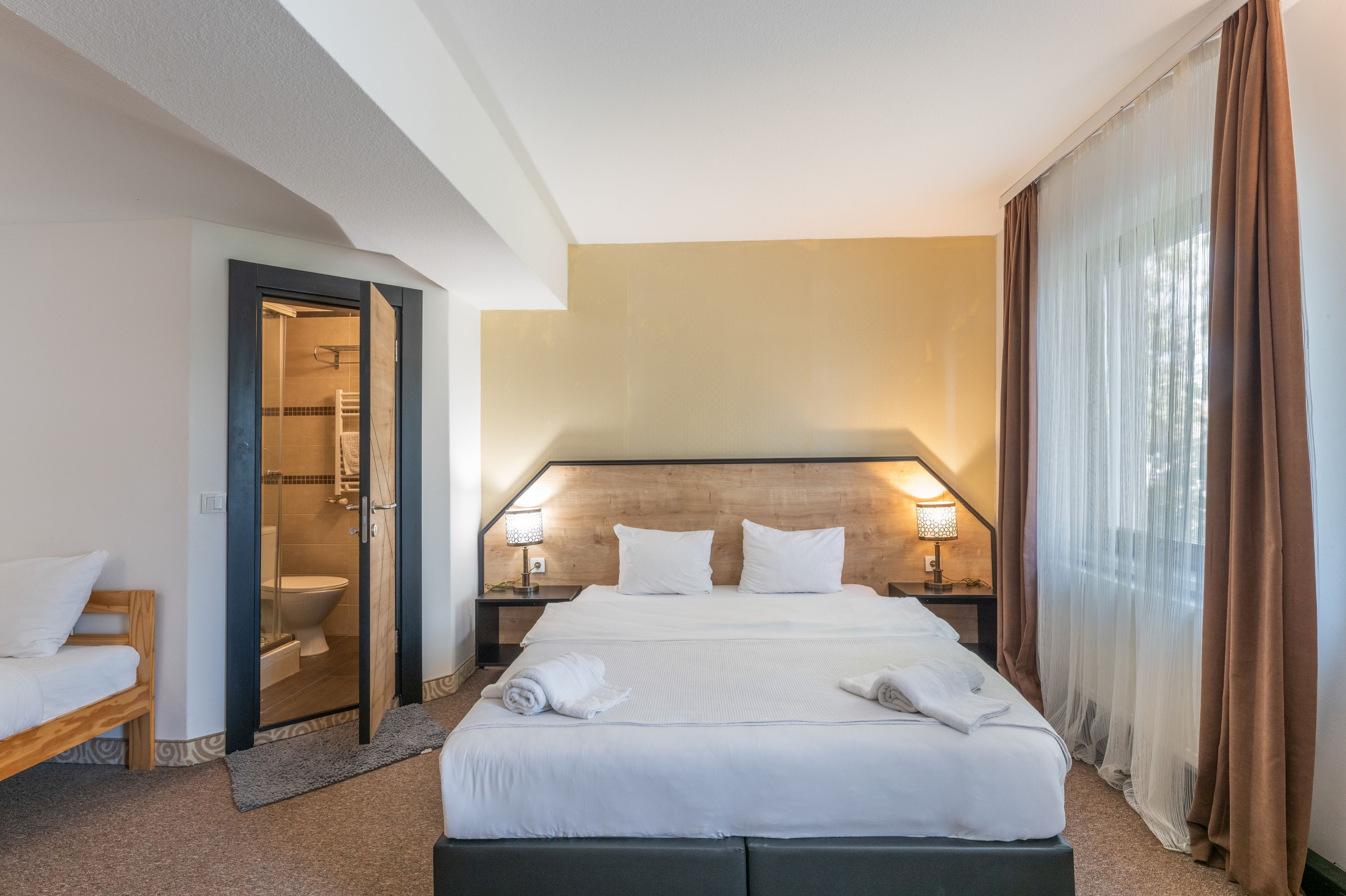 A hotel room with a bed, two bedside lamps, a wooden headboard, a bathroom with a glass door, and a window with curtains.