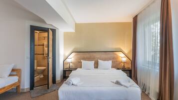 A hotel room with a bed, two bedside lamps, a wooden headboard, a bathroom with a glass door, and a window with curtains.