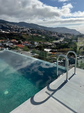 Ein Pool ohne Ende mit klarem Wasser, Metallgeländern und Blick auf eine Stadt und Berge im Hintergrund.