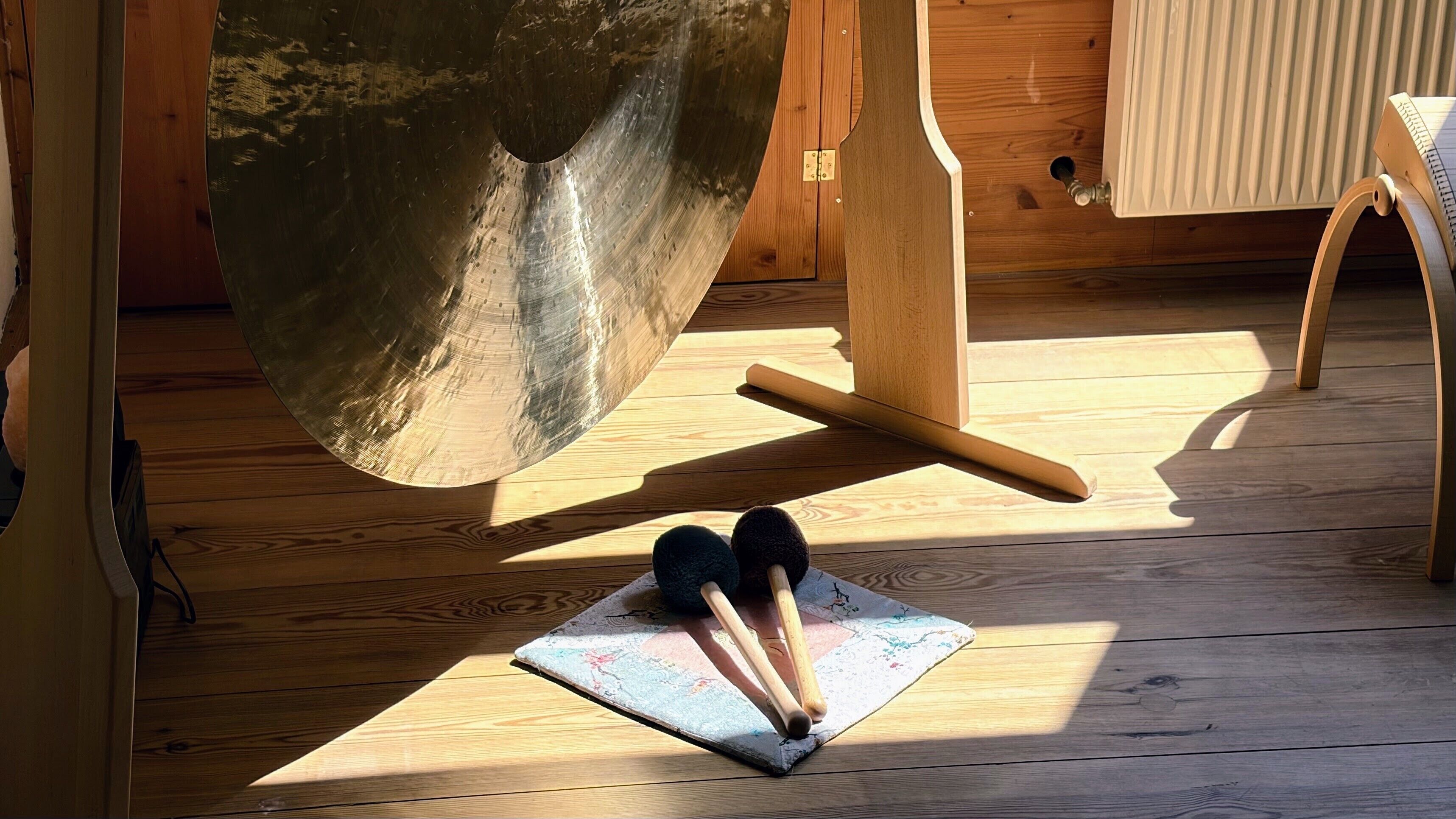 A wooden floor with a large gong, a wooden stand, and a pair of drumsticks on a mat.