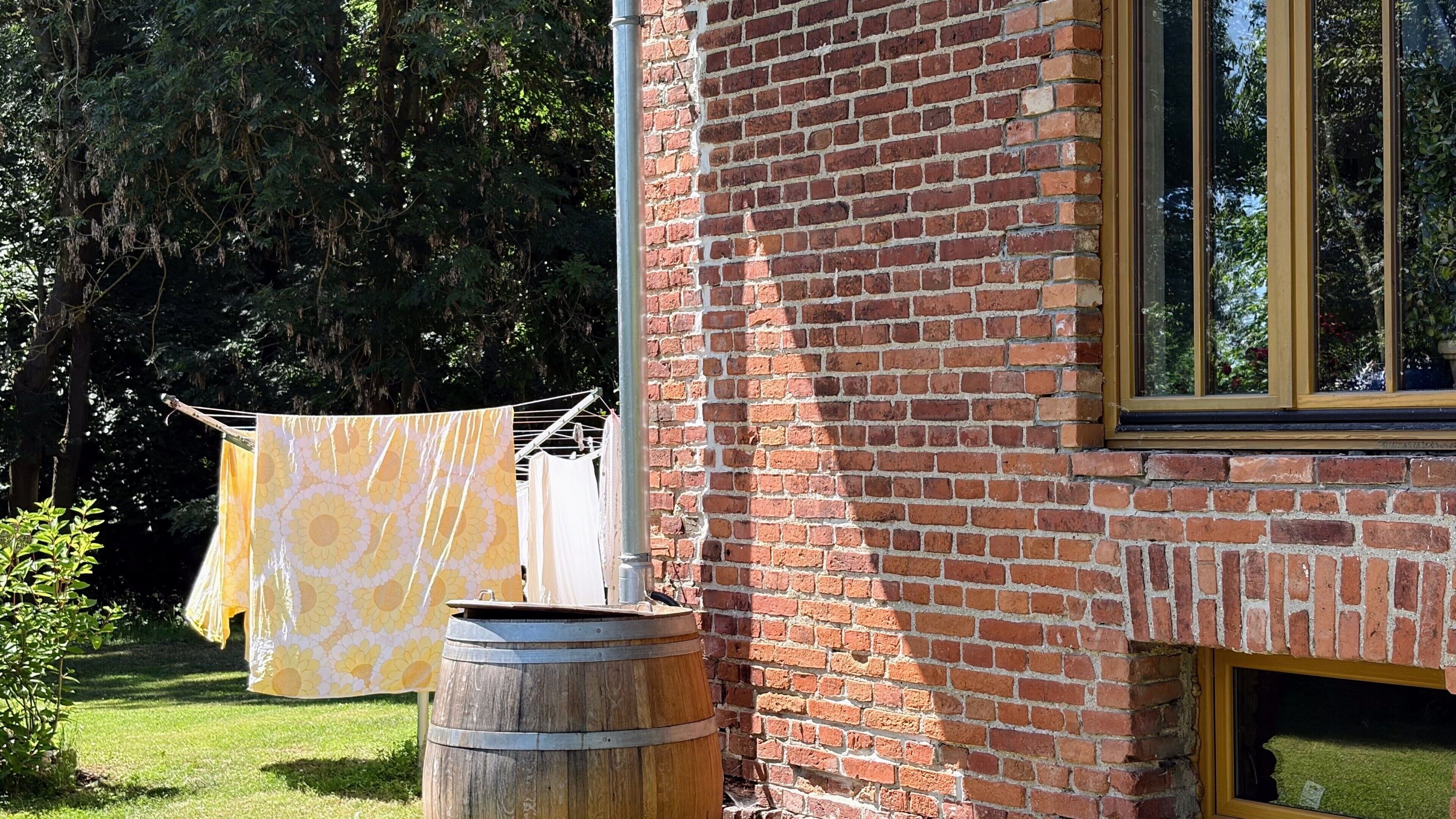 Un bâtiment en briques avec une fenêtre, une corde à linge sur laquelle est suspendu un tissu jaune à motifs, et un tonneau en bois.