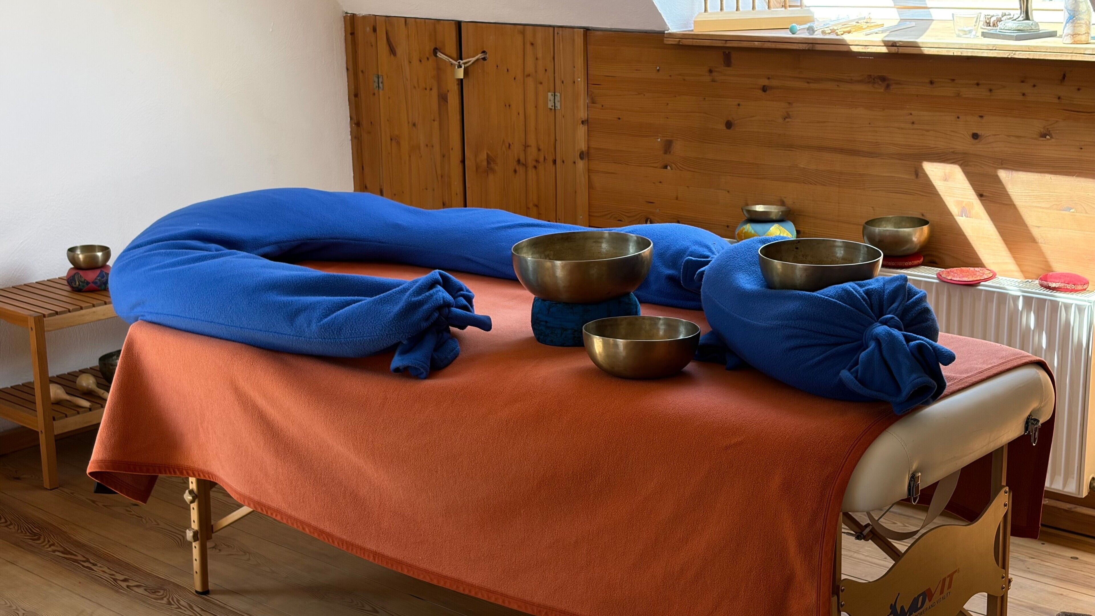 Uma mesa de massagem com uma toalha azul e uma toalha vermelha, rodeada por tigelas de metal e um armário de madeira.