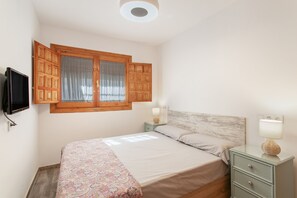 Et soveværelse med en seng, et natbord, en lampe, et fjernsyn ophængt på væggen og et vindue med træpersienner.