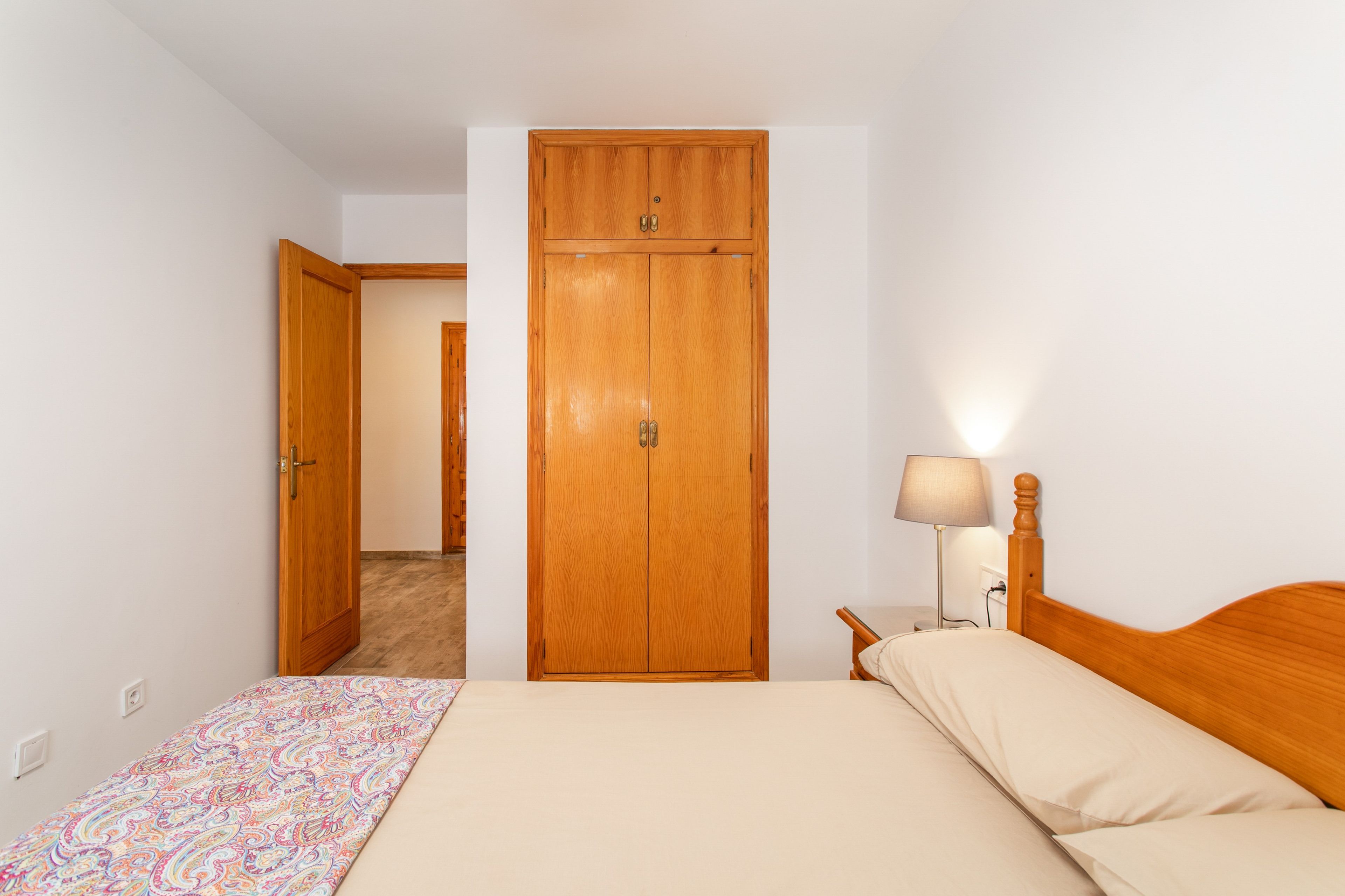 Een slaapkamer met een bed, een houten kledingkast, een nachtkastje met een lamp, en een deur die naar een andere kamer leidt.