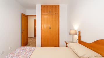 Een slaapkamer met een bed, een houten kledingkast, een nachtkastje met een lamp, en een deur die naar een andere kamer leidt.