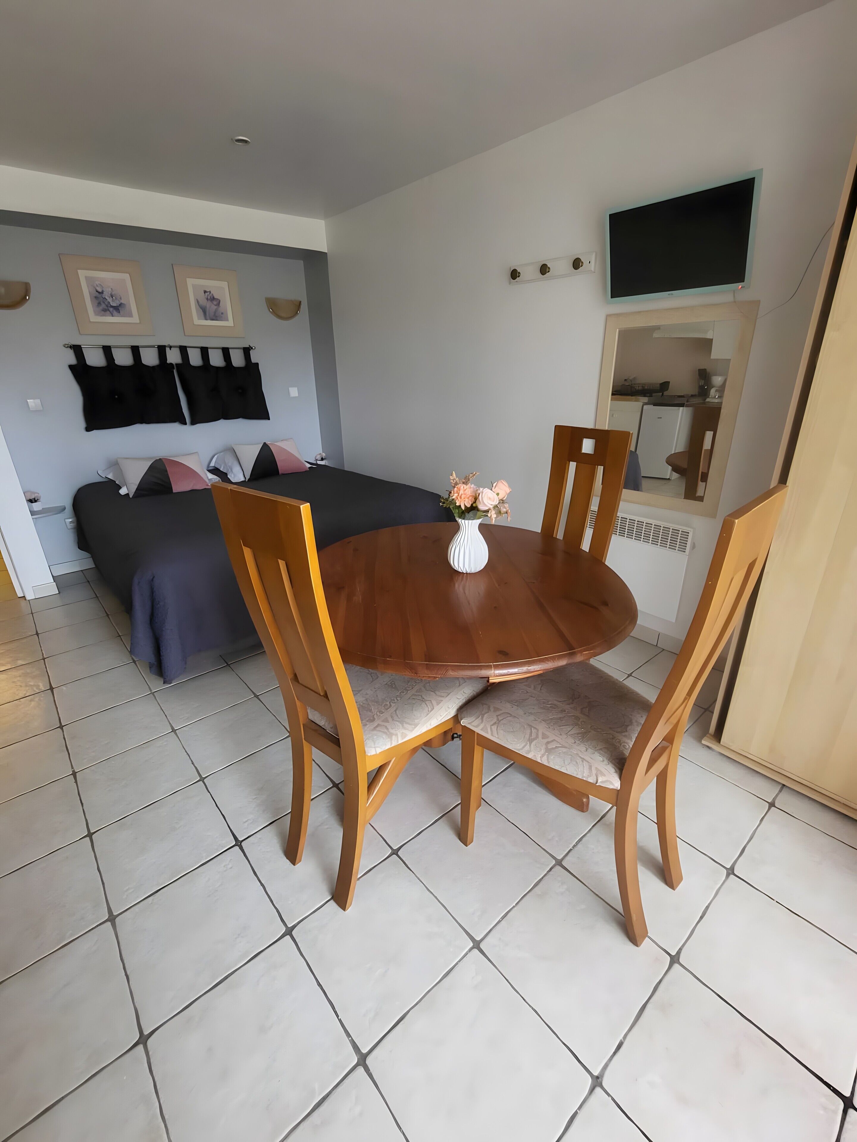 A room with a bed, a dining table with chairs, a small white vase with flowers, and a television mounted on the wall.