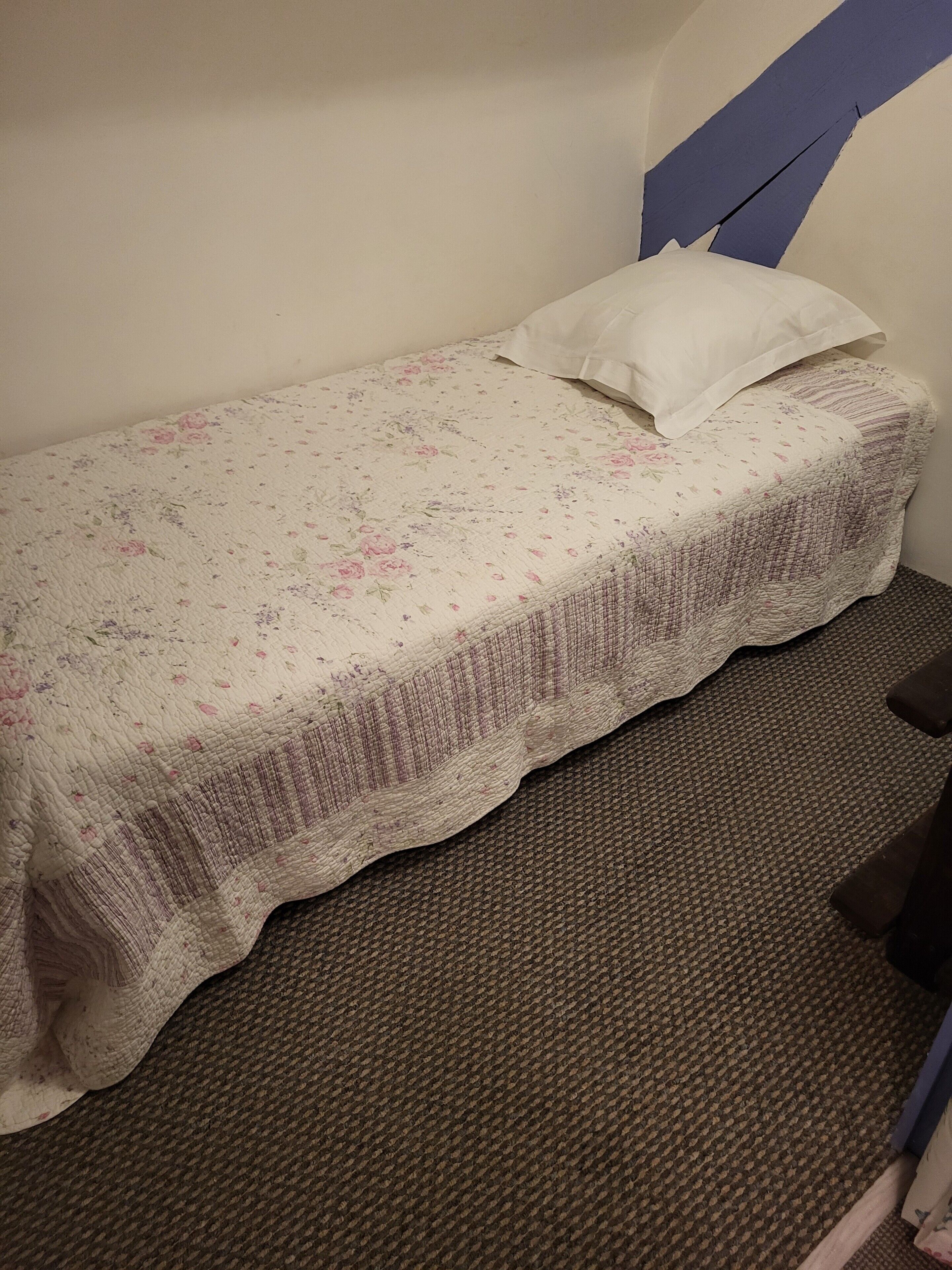 Ein kleines Zimmer mit einem Einzelbett, einer Bettdecke mit Blumenmuster, einem weißen Kopfkissen und einem braunen Teppichboden.
