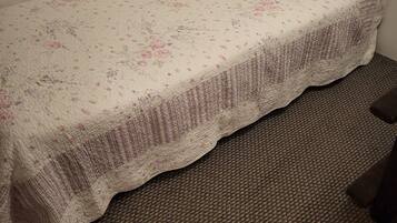 Ein kleines Zimmer mit einem Einzelbett, einer Bettdecke mit Blumenmuster, einem weißen Kopfkissen und einem braunen Teppichboden.