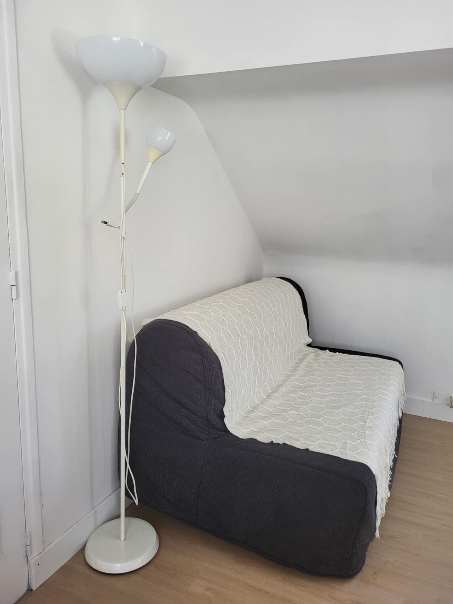 Ein kleines Zimmer mit einem Einzelbett, einer weißen Stehlampe und einem dunkelgrauen Sofa.